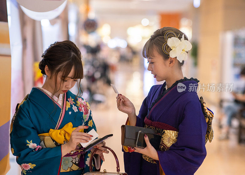 年轻女性穿着“Furisode”和服，看着印在“Purikura”照片贴纸摊位上的“Seijin Shiki”成人仪式贴纸